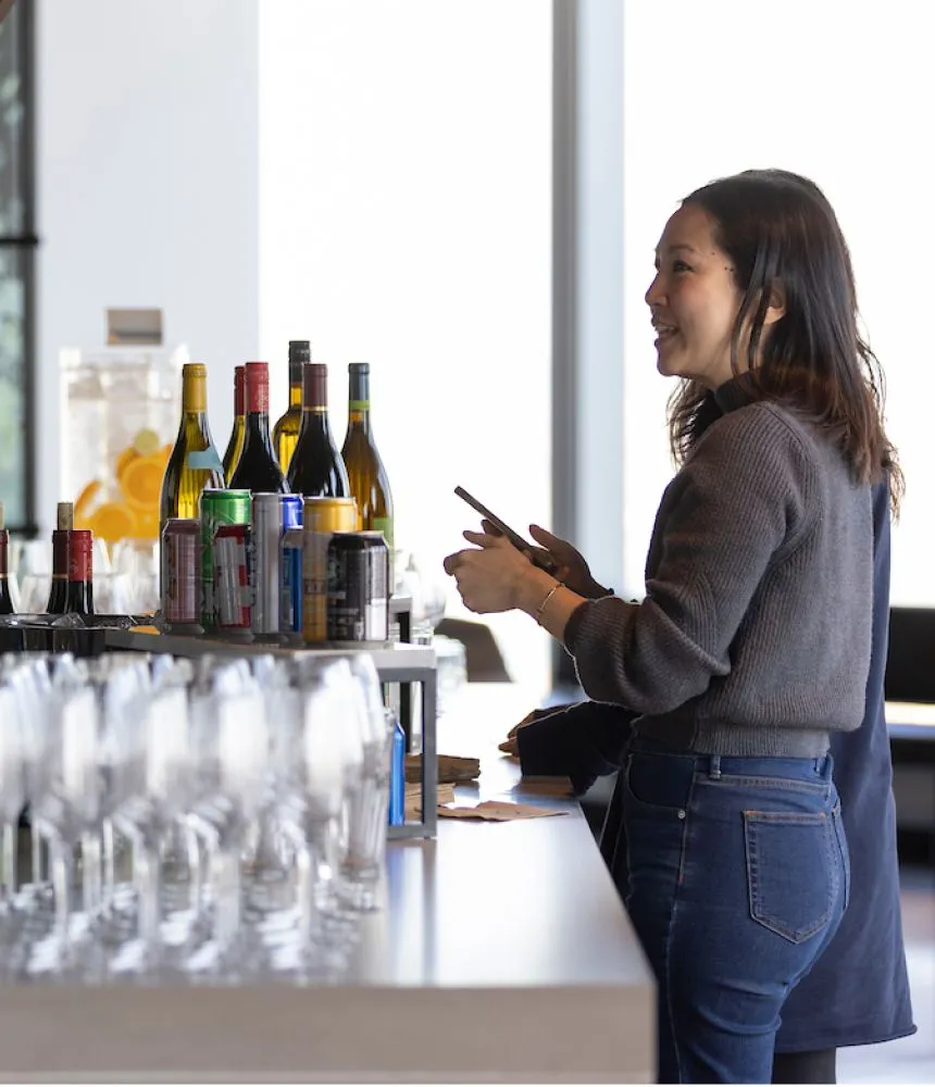 A woman standing at a bar filled with champagne glasses and wine bottles at an corporate event at Well& by Durst at One World Trade Center..