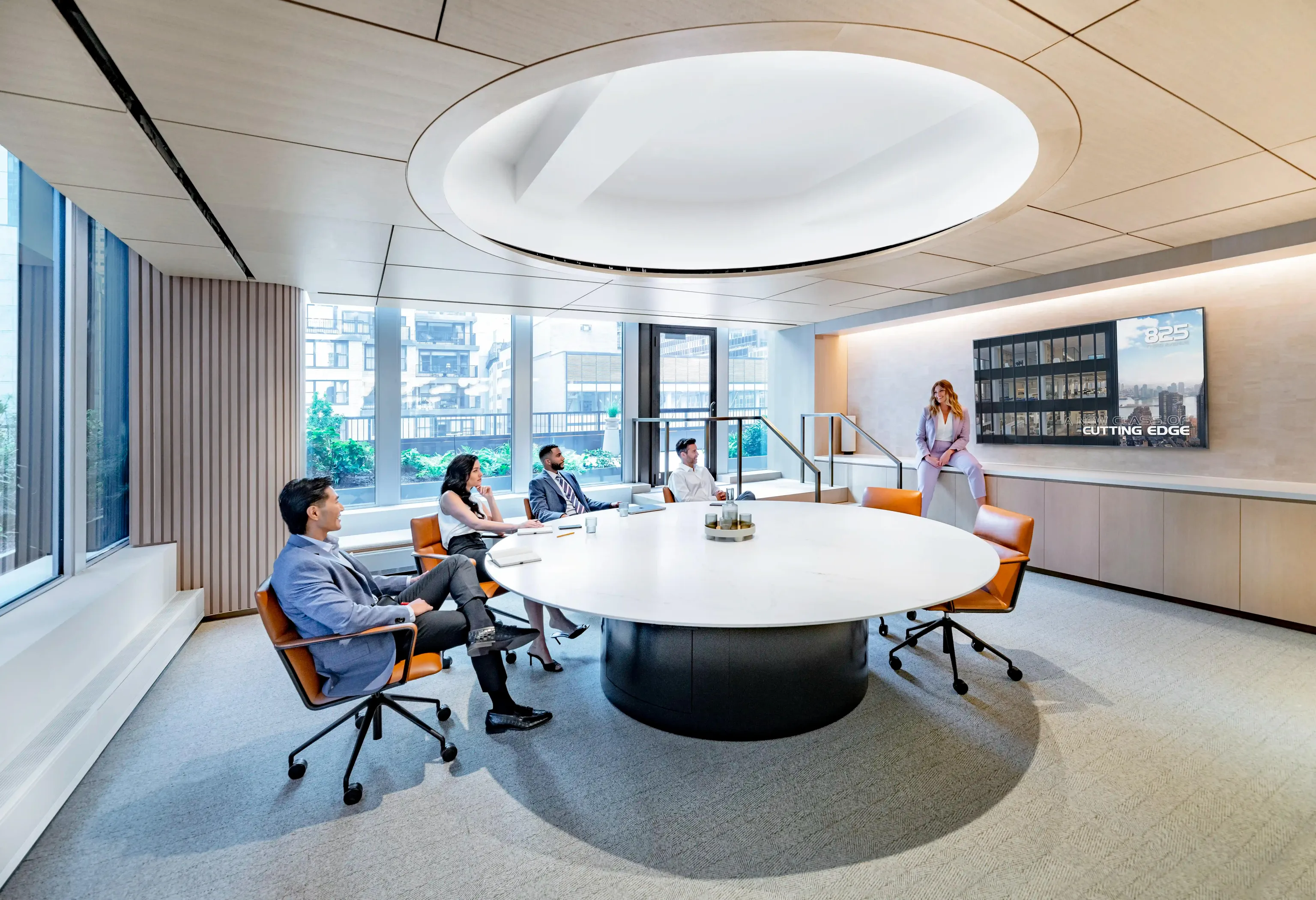 A group of diverse individuals sitting in a very modern looking conference room engaged in active discussion. A well-dressed female is sitting to the side next to the tv as the center of attention.