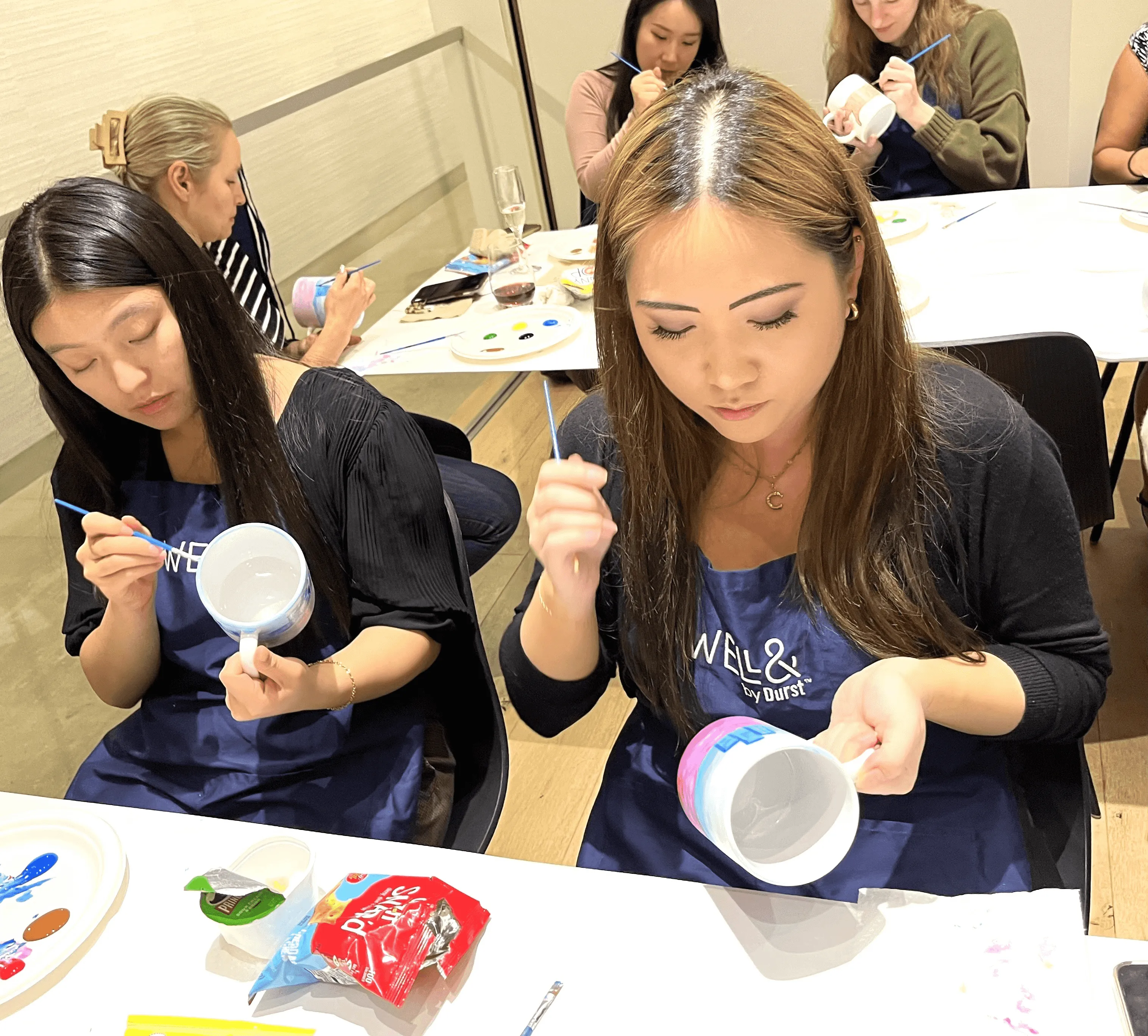 A diverse group of women painting mugs with colorful brushes. They are all wearing "Well& by Durst" aprons.