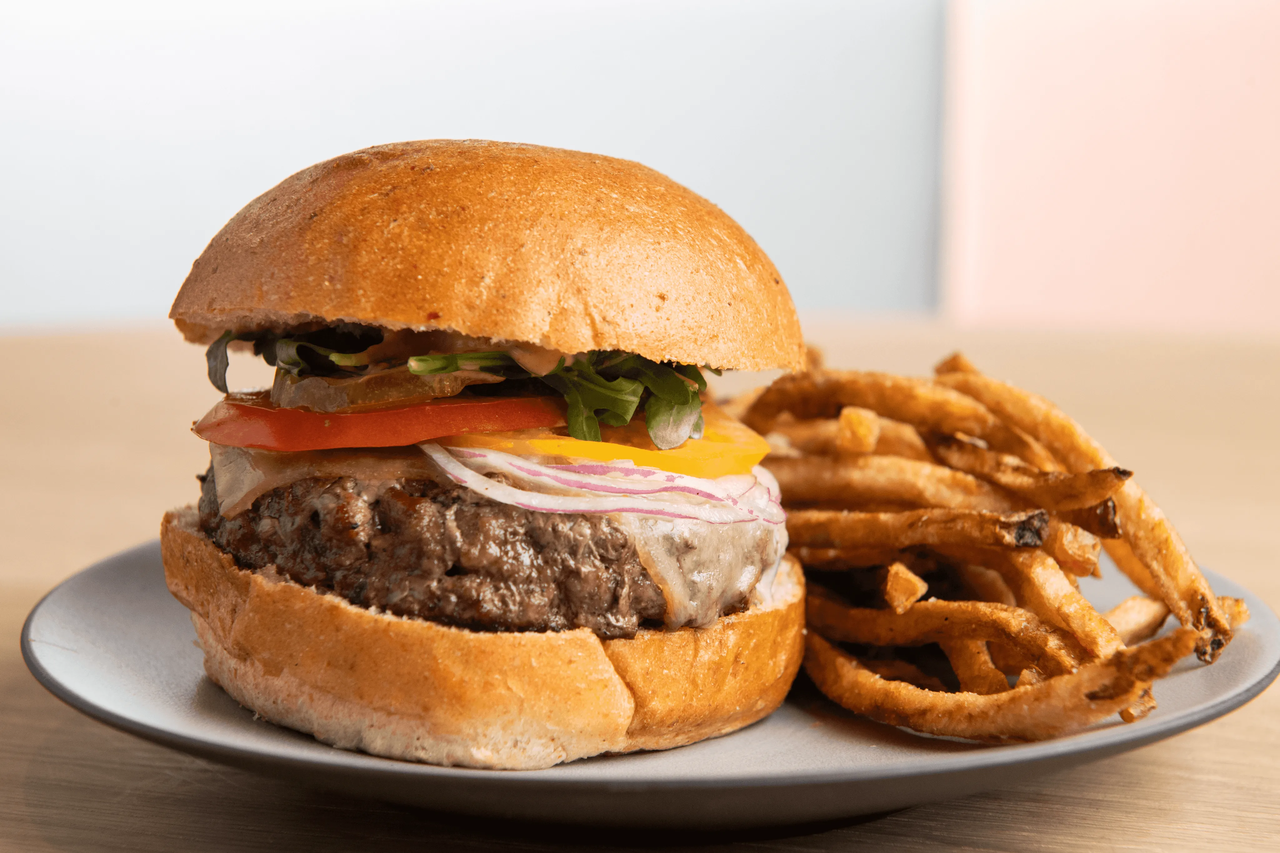 A juicy hamburger with a sesame seed bun, topped with lettuce, tomato, cheese, and a beef patty, sits on a white plate next to a generous serving of crispy, golden-brown French fries.