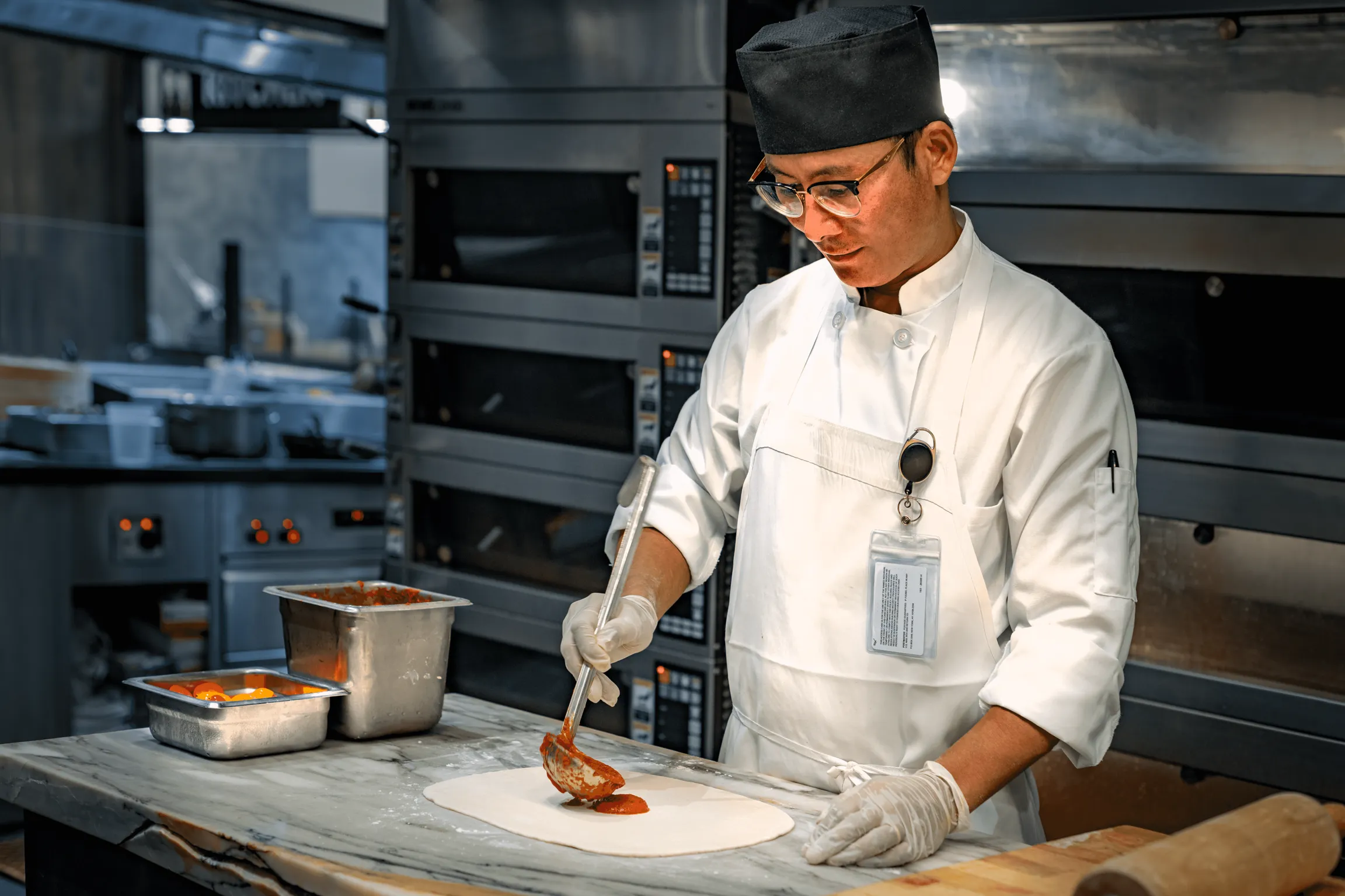 A pizza chef in a kitchen covering pizza dough with garnishes and sauces.