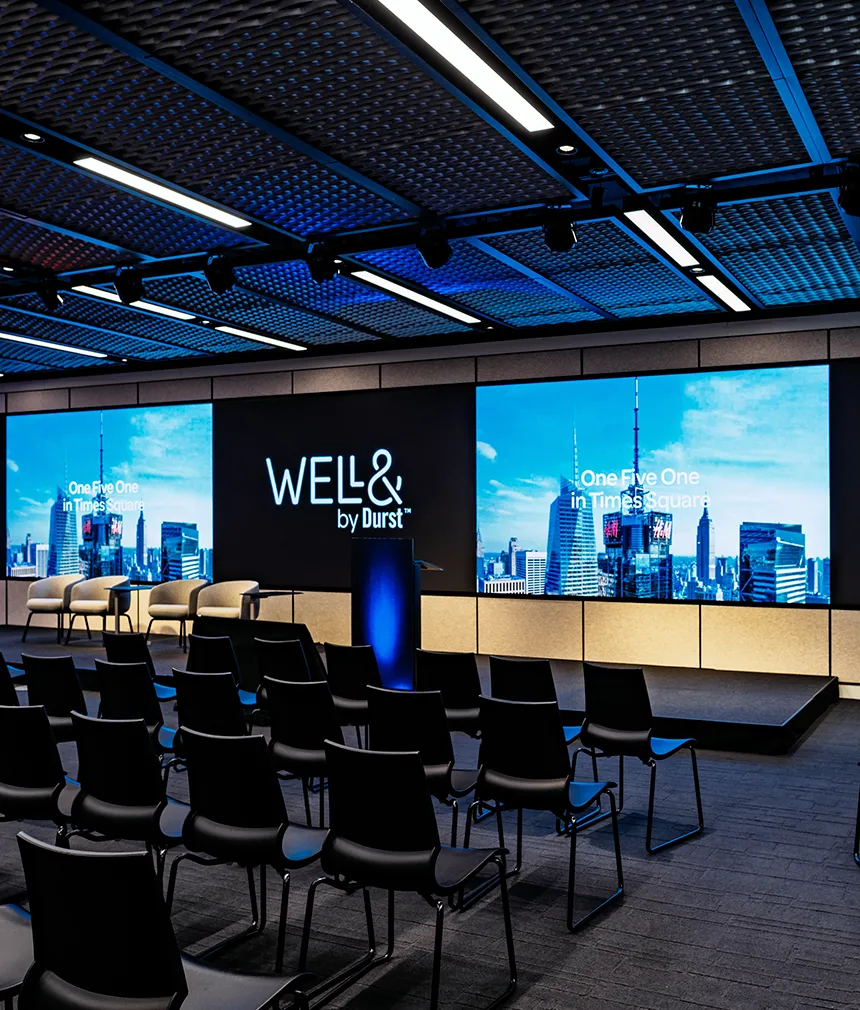 151 West 42nd conference space filled with empty chairs and a large screen that extends the far end of a wall. It displays the "Well& by Durst" logo on it.