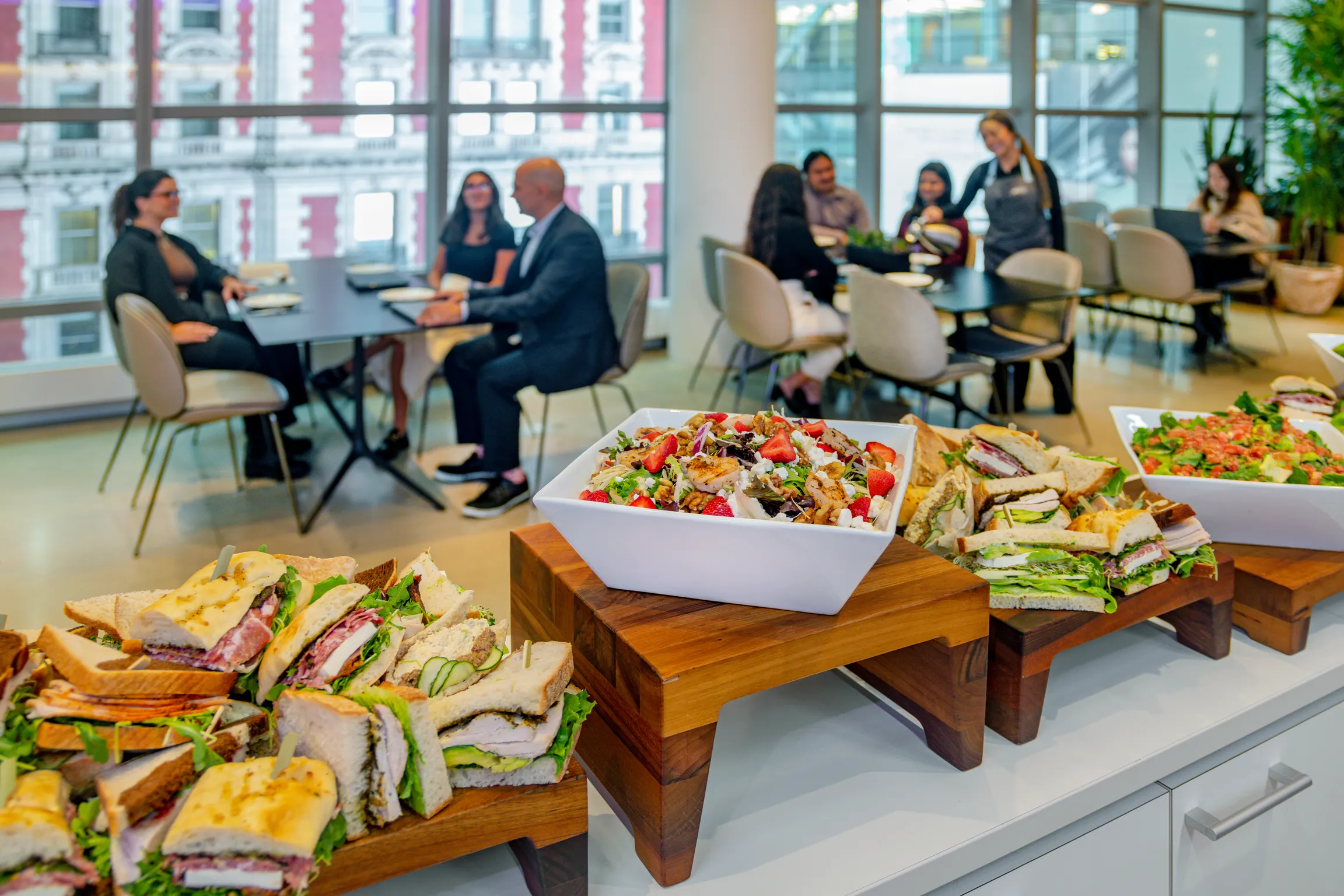 People sitting at tables with sandwiches and salads, with a catering bar and people eating in the background.