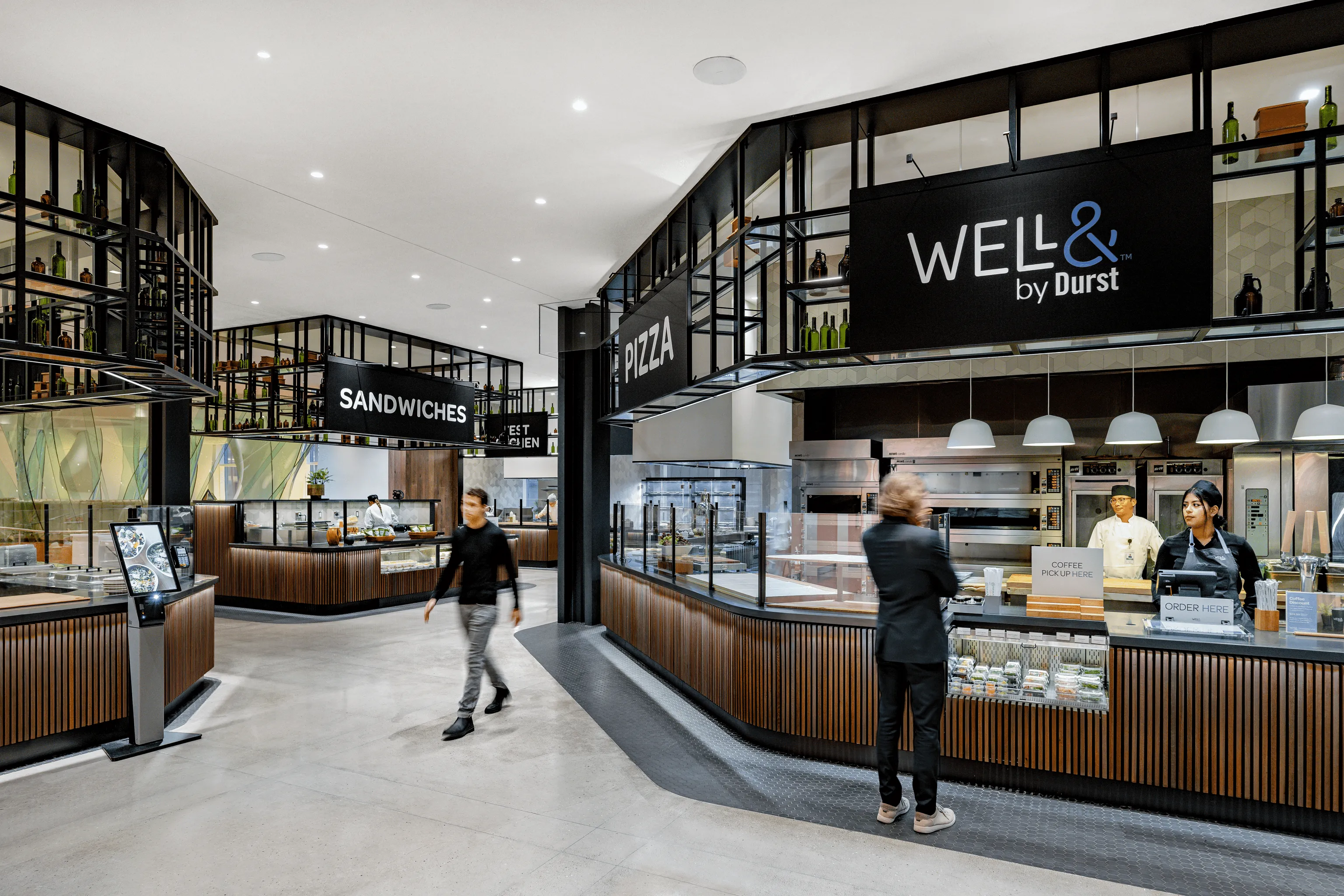 Modern cafeteria interior with a sign displaying "Well& by Durst", featuring a sandwich bar, juice station, and seating area under soft lighting, with staff serving customers.