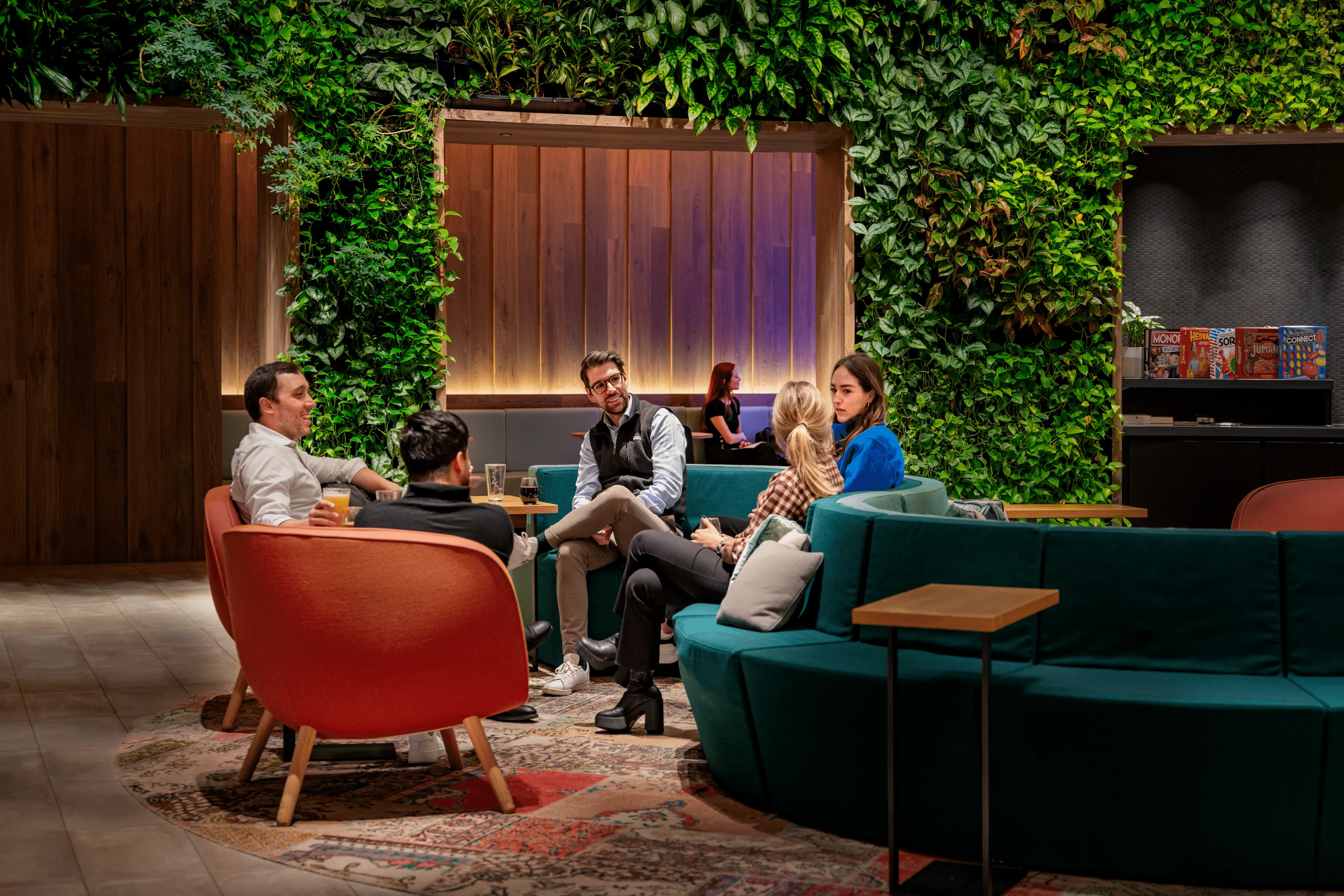 "The Green" at 151 West 42nd—A modern lounge area with lush green plant walls. Five people are seated in a relaxed setting on comfortable chairs and sofas, engaged in conversation.