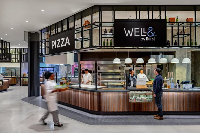 Modern cafeteria interior with a sign displaying "Well& by Durst", featuring a sandwich bar, juice station, and seating area under soft lighting, with staff serving customers.